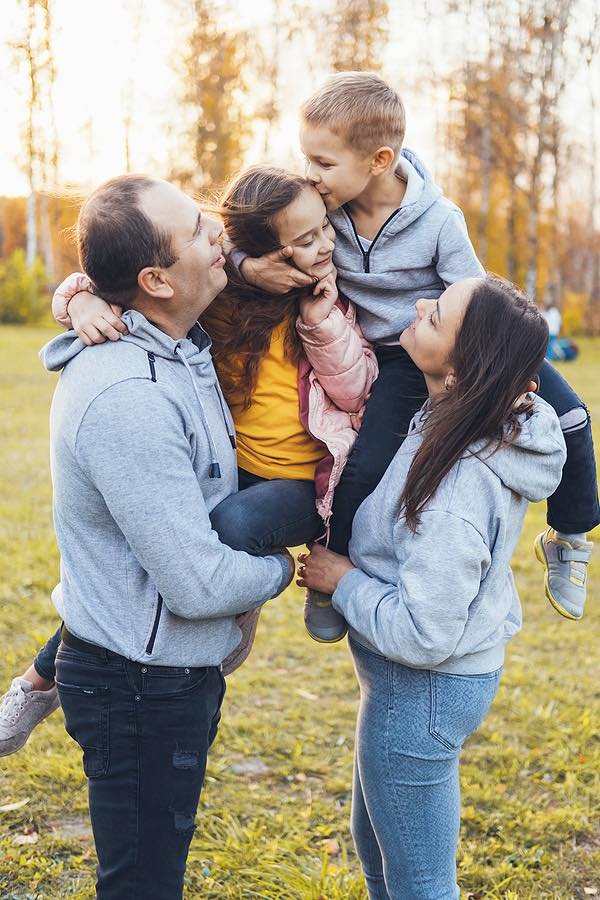 A Family Of Four Happily Spending Their Free Time In The Park, The Brother Kissing His Sister On The